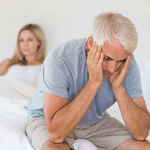 A worried older man sitting on the edge of a bed with his partner behind him